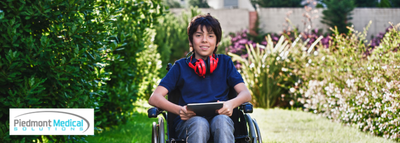 Boy in wheelchair outside smiling with headphones around his neck and tablet in his hands 