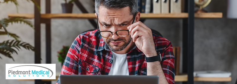 Man holding the frame of his glasses while looking down at his laptop screen confused