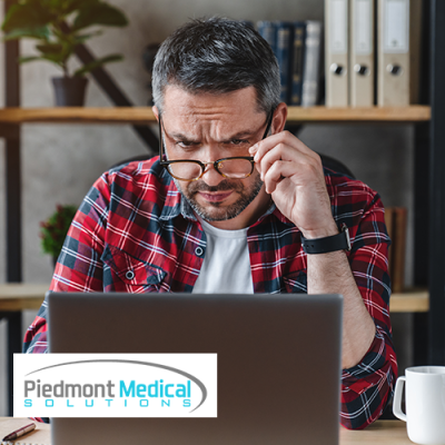 Man holding the frame of his glasses while looking down at his laptop screen confused